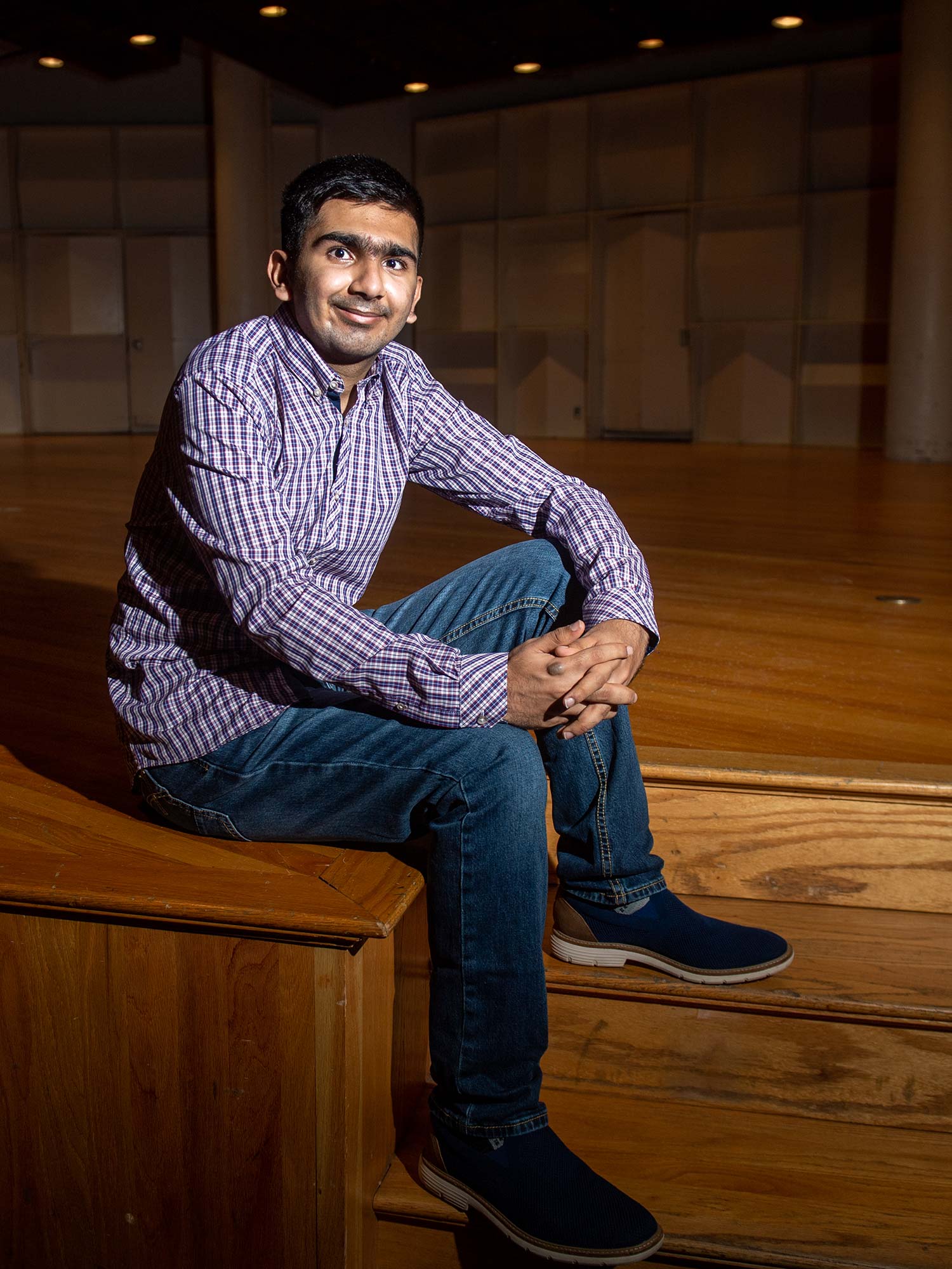 Photo of Faizaan Firoz (Pardee’25) sitting on the stairs leading up to a stage with a checkered button down and jeans on. He smiles and the dramatic lighting casts a long shadow on the stage behind him.