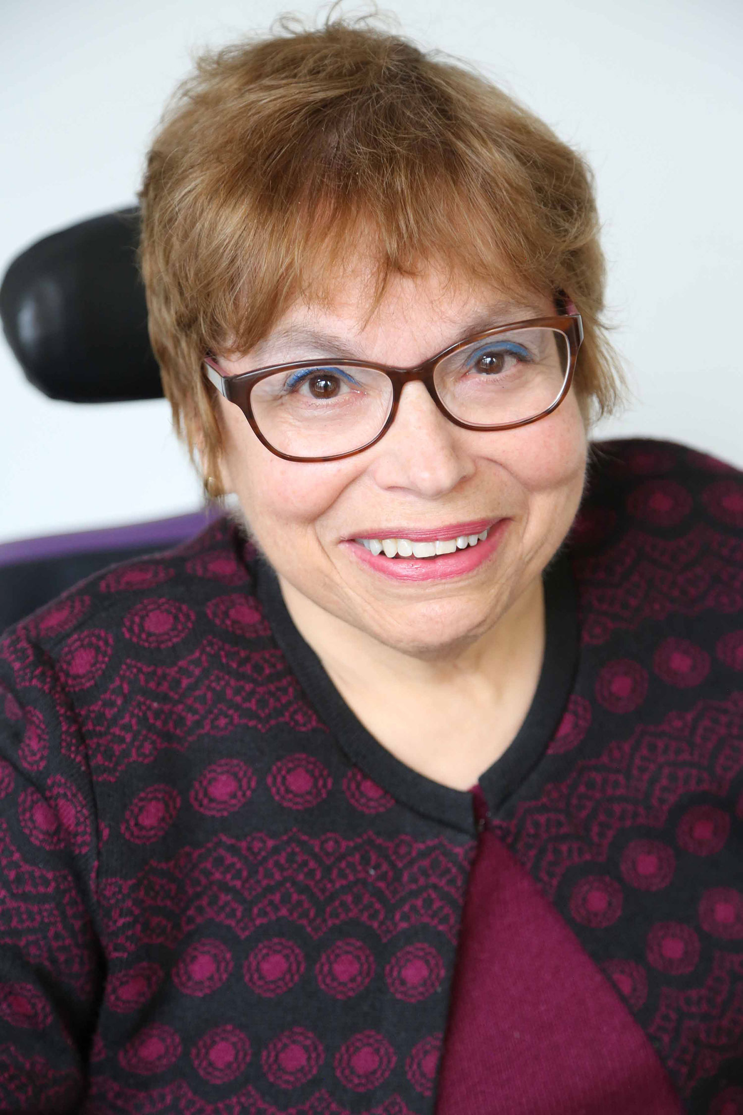 Headshot of Judith Heumann, smiling. She wears purple-ish glasses that match with her sweater, which has a geometric pattern. Her hair is goldish; Judith is white and is a wheelchair user.