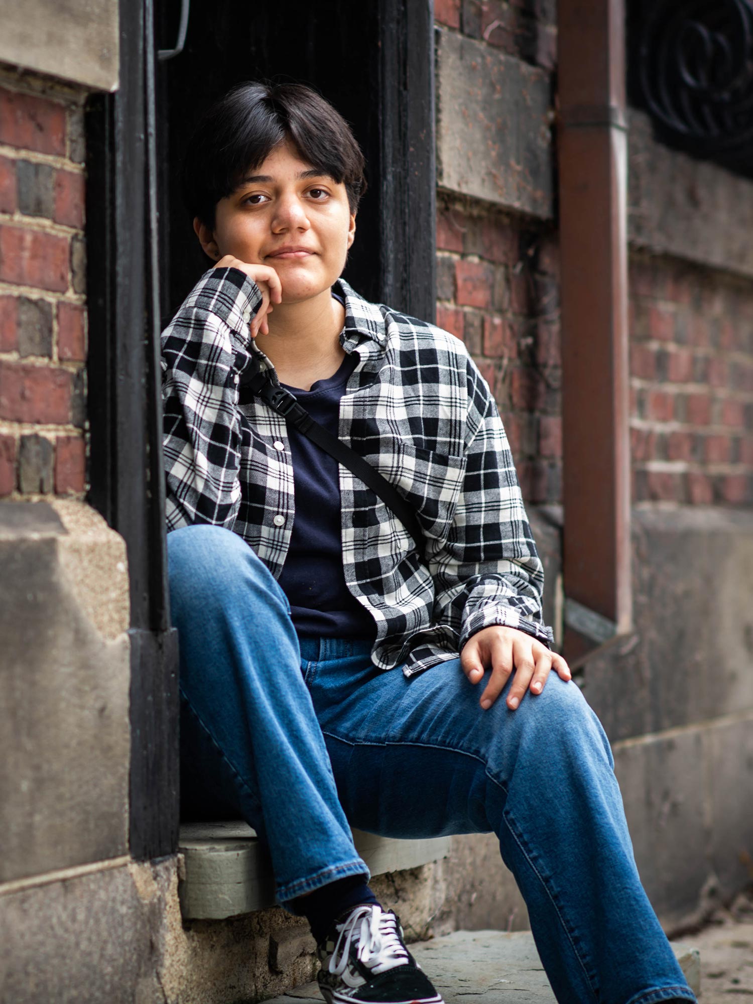 Photo of Ash Vasquez (CFA’25), sitting on a step between two brick walls. His hand is on his face, and he wears a plaid shirt and jeans.