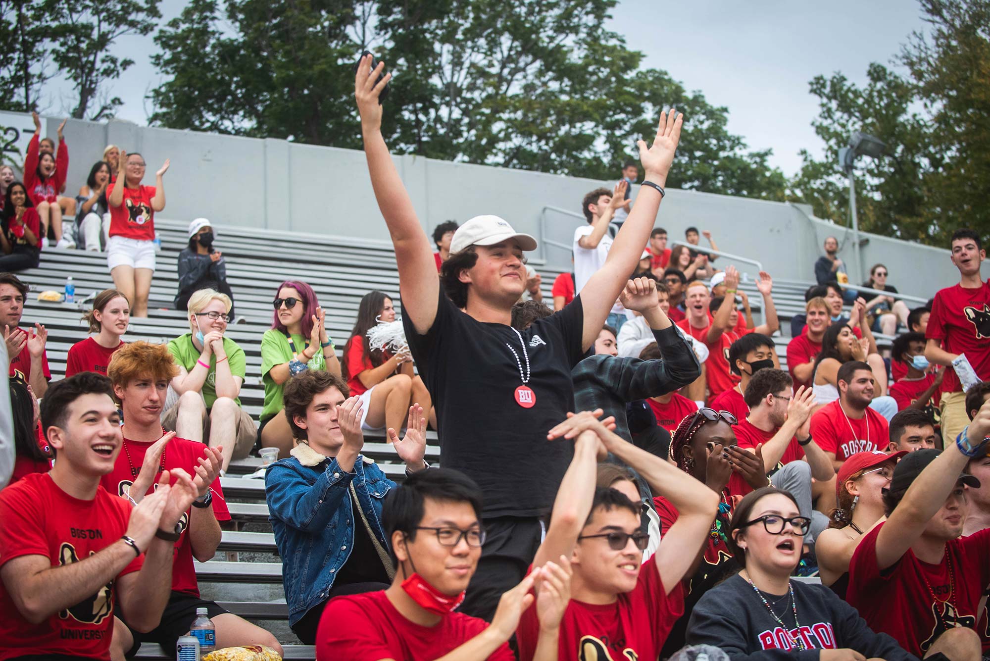 Nathan Basman (QST 25) celebrates a goal during the woman’s soccer game at the Terrier Tailgate on Sunday, August 29, 2021. He extends his arms above his head, and wears a white baseball cap. Students, many dressed in red, sitting on the bleachers around him.