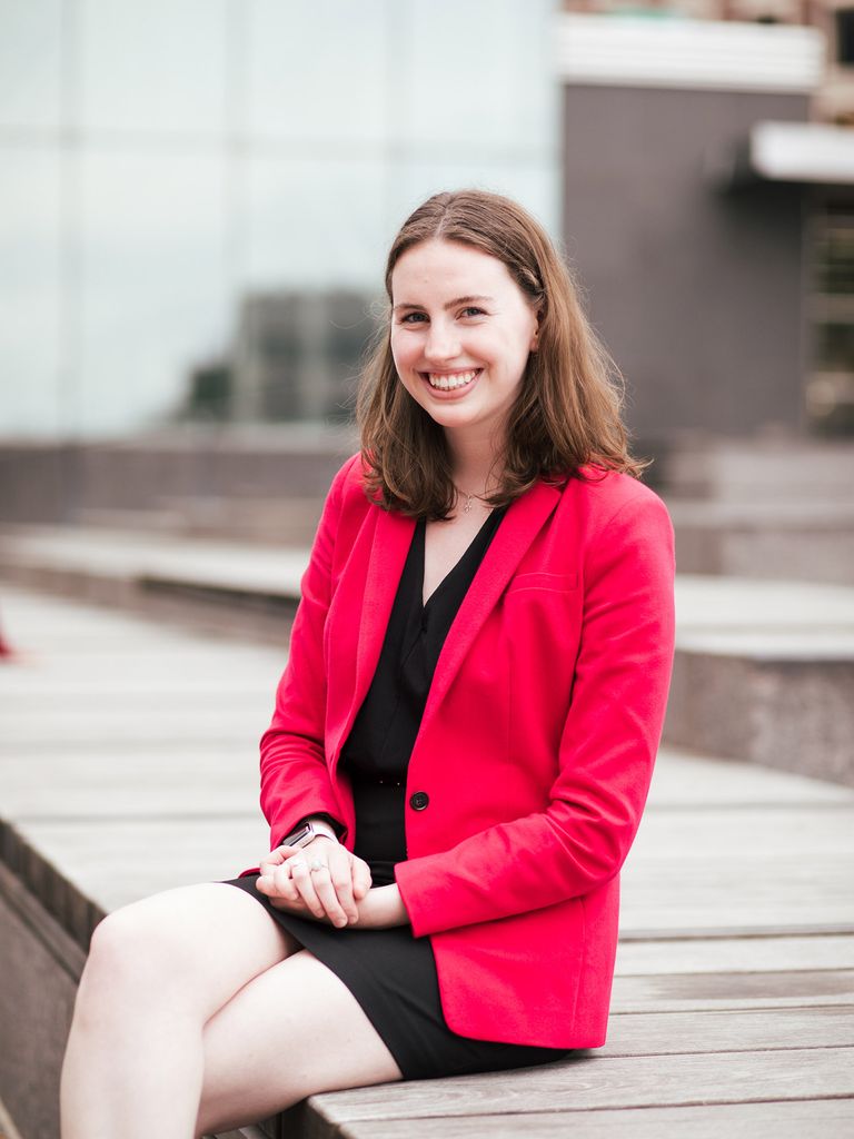 Photo of Lily Kelly (CAS’22) with a bright pink blazer and black dress on sitting on what appears to be a wooden bleacher seat in an outdoor auditorium.