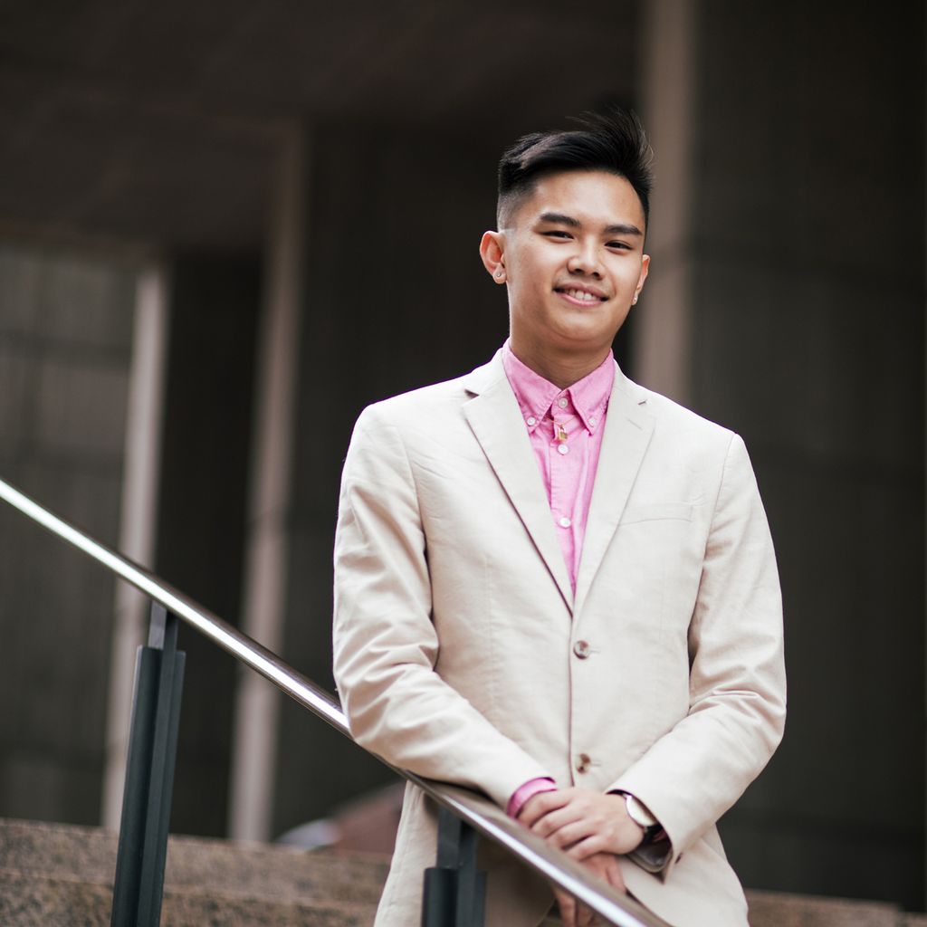 Photo of Avi Nguyen (CAS’22) in a beige suit with a pink button down. He leans along the hand rail of stairs outside at government center and smiles.