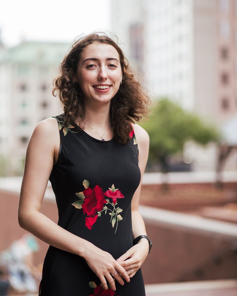 Photo of Julia Sullivan (Sargent’22) standing in government center. She wears a black dress with a red floral pattern and smiles with her hands partially clasped.