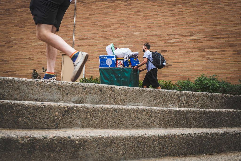 Photo taken of a student pushing a green bin filled with their belongings from ground level; a few stairs are seen in the foreground.