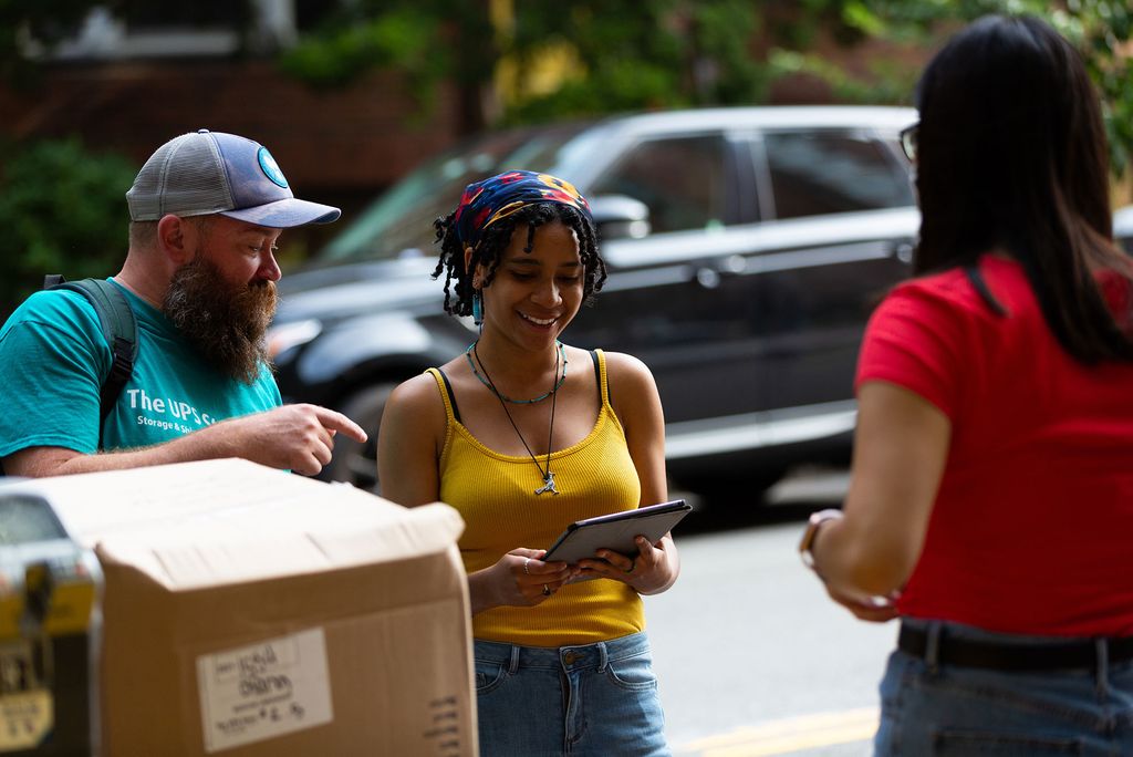 Someone who looks like an RA, smiles, and helps people who are moving in; they hold a tablet of some kind.