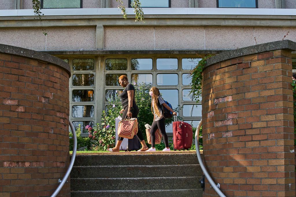 Photo of a student and her mom walking near the towers rolling suitcases, foreground by brick walls and a flight of stairs.