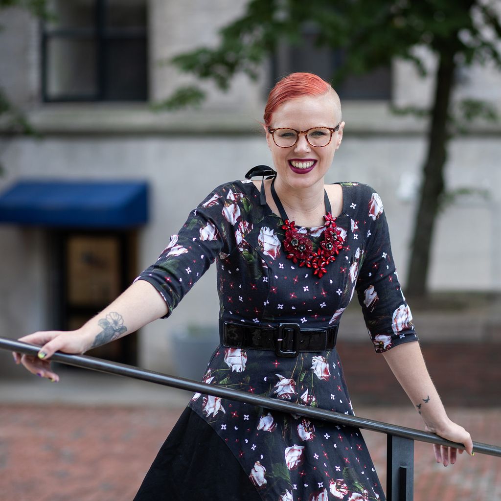 Photo Kimberly M. Rhoten (GRS’26) leaning against a hand rail somewhere in government center. She wears a black dress with a red and white floral pattern and a chunky red necklace.