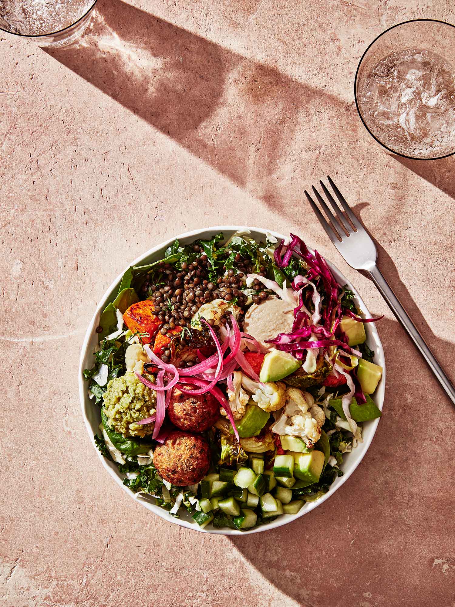 Aerial photo of a grain bowl with what looks like falafel. The bowl is stacked high with greens, onions, cabbage, etc and rests on a pink surface.