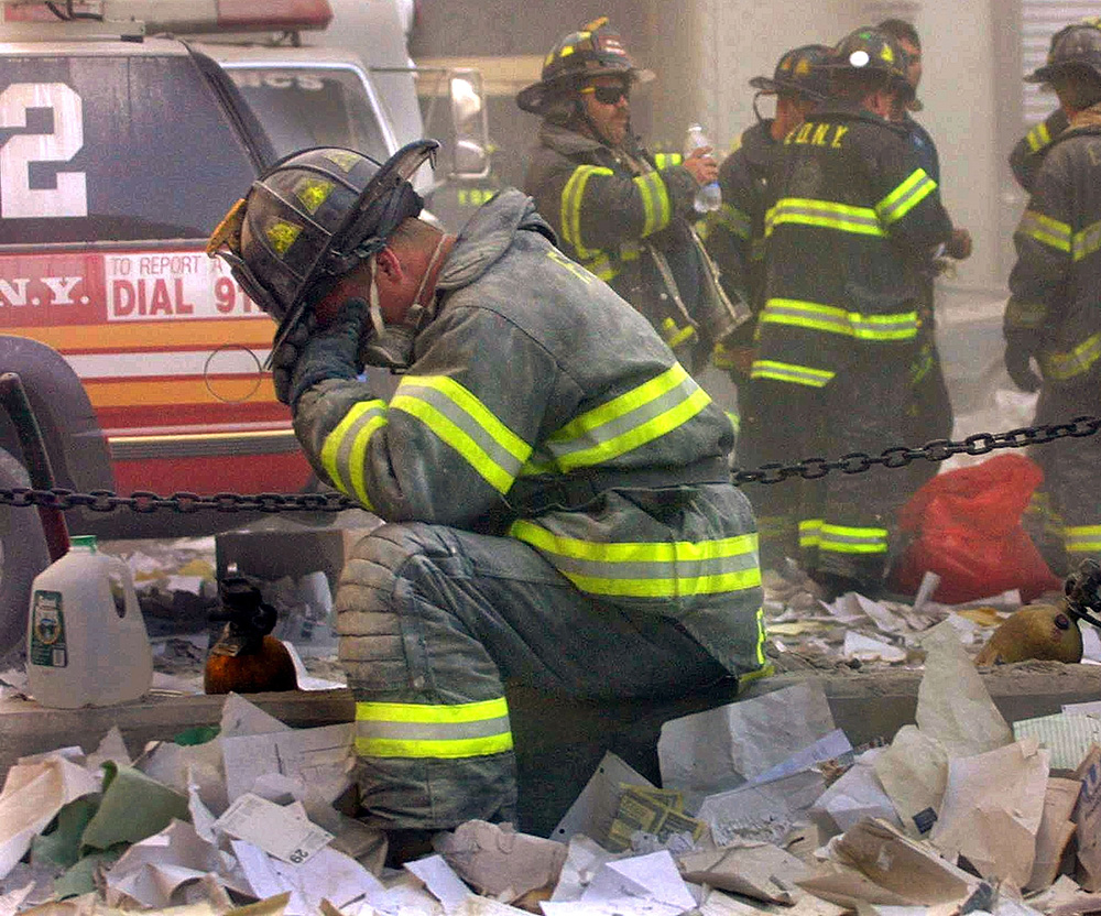 Photo of frefighter Gerard McGibbon, kneeling, with his face in his hands, praying after the World Trade Center buildings collapsed. A group of firefighters behind him are seen hydrating. Rubble is all around them and the air is ashy.