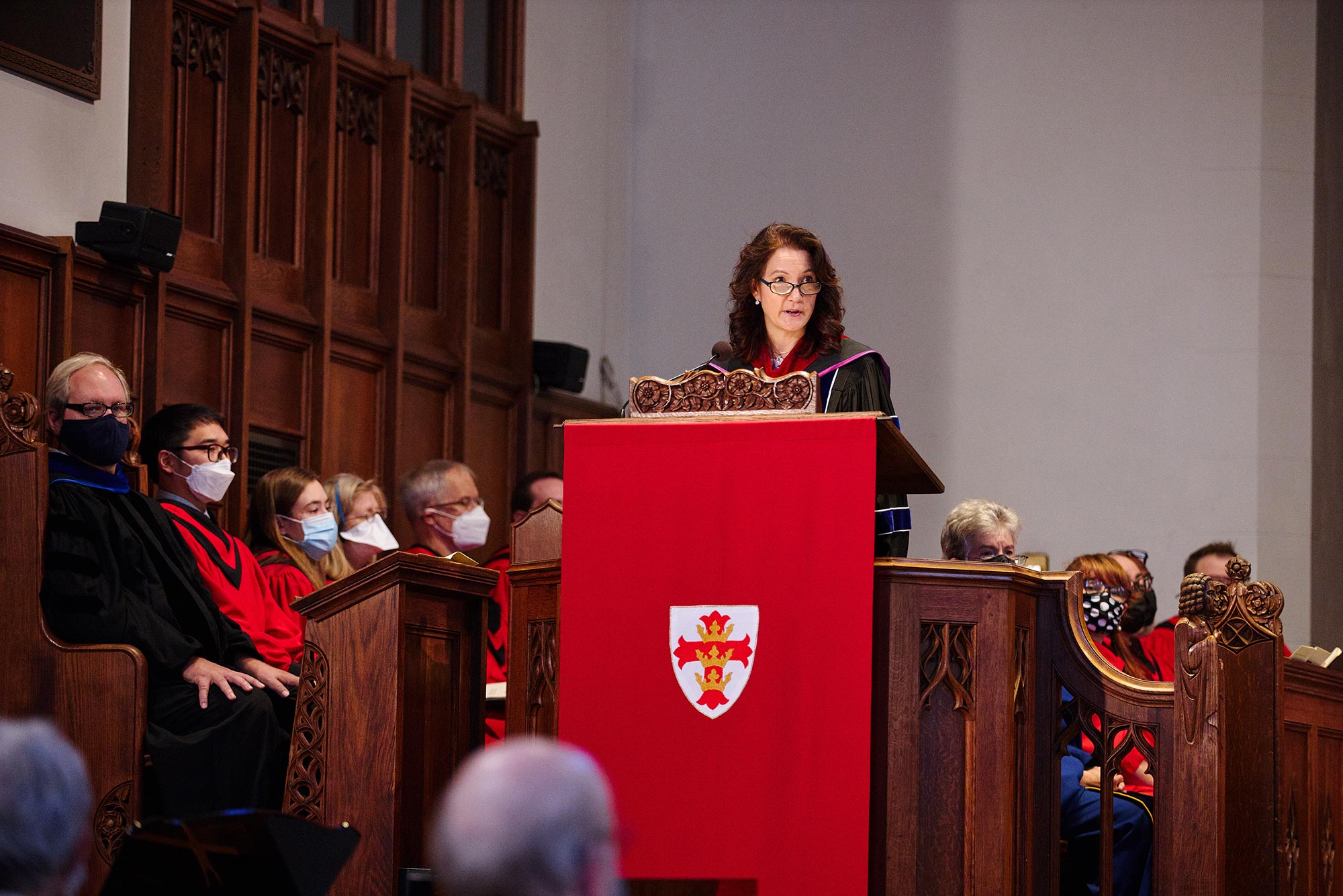 Photo of G. Sujin Pak’s dean installation service at the school of theology. Pak stands at a podium covered with a bright red flag and looks off to her left into the congregation, presumably she is speaking. Other members of the service sit behind her in a large wooden pew.