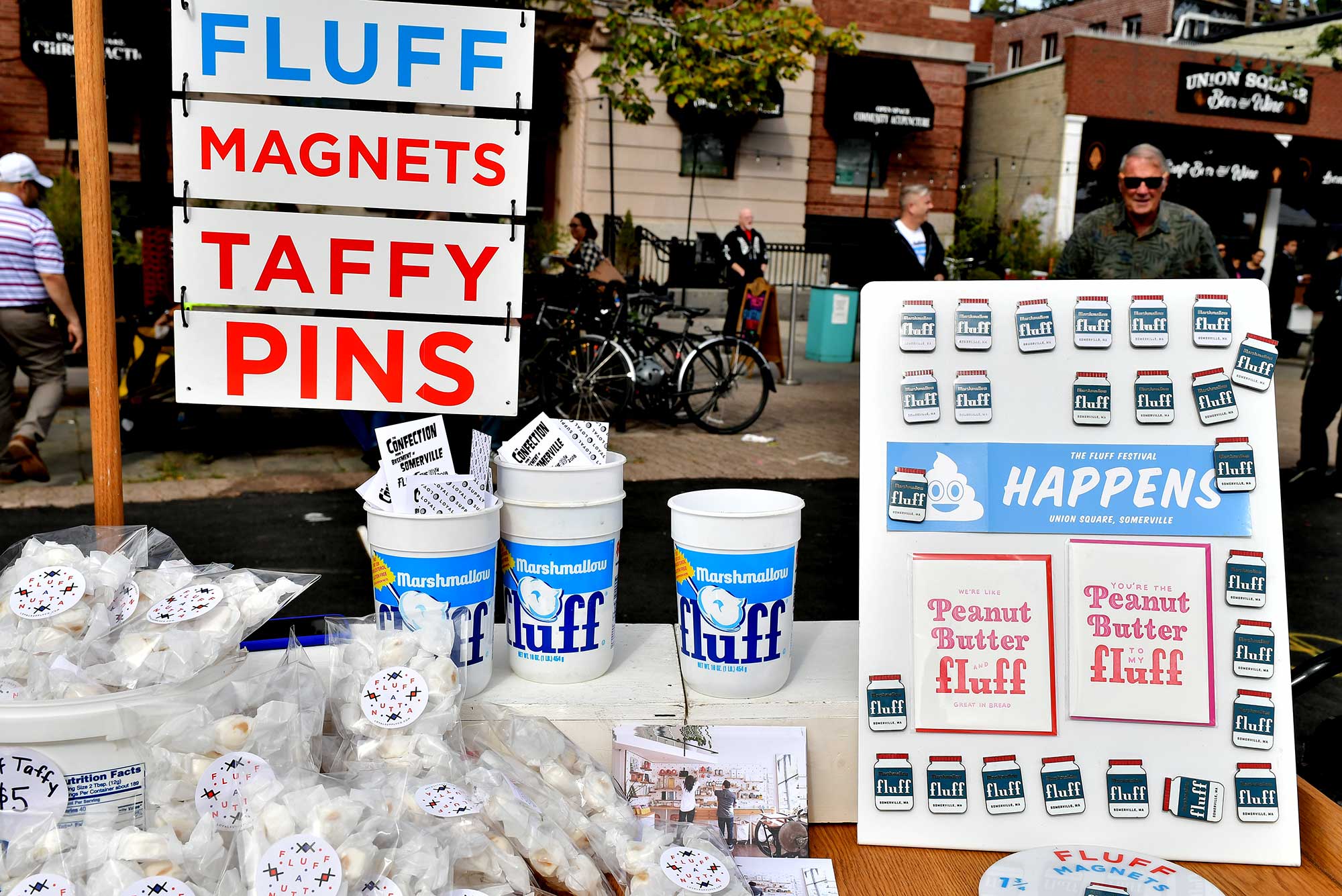 Photo of a table filled with taffy and fluff containers filled with business cards, as well as a display of pins in the shape of jars of fluff. Behind the table, a sign reads “FLUFF. MAGNETS. TAFFY. PINS.” Photo was taken at What the Fluff Festival on September 22, 2018 in Somerville, Massachusetts.