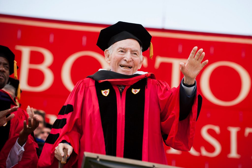 Photo of George Wein (CAS'50), wearing a red robe and black hat, raising his left hand, and using a cane in the other, as he is presented with an honorary degree during the 2015 Boston University Commencement at Nickerson Field on May 17, 2015. He smiles, and the Boston University sign is seen behind him.