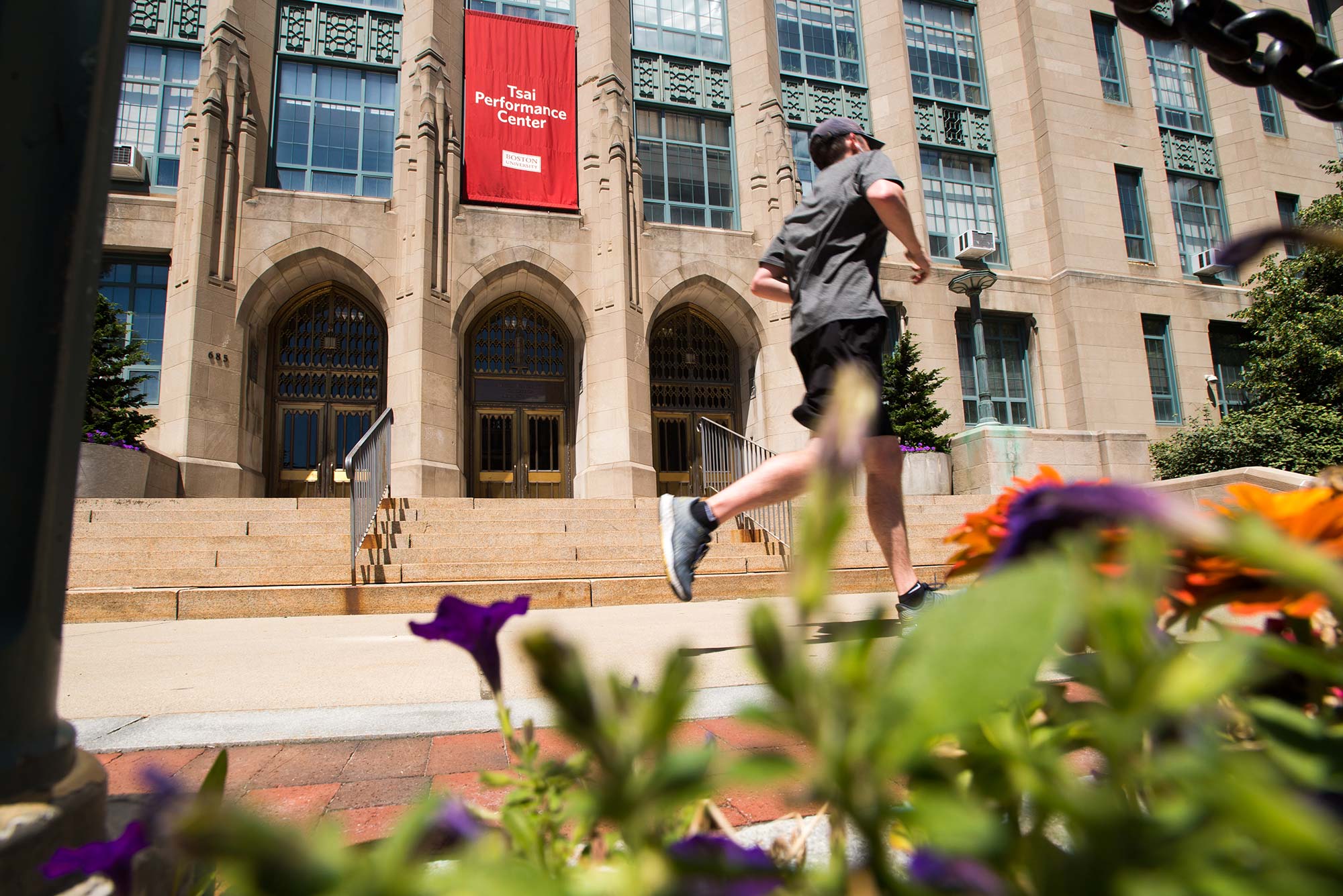 Photo taken at street level on Comm Ave towards the sidewalk and the Tsai Performance Center, where a runner in a gray shirt and black shorts runs by. Flowers are seen in the foreground.