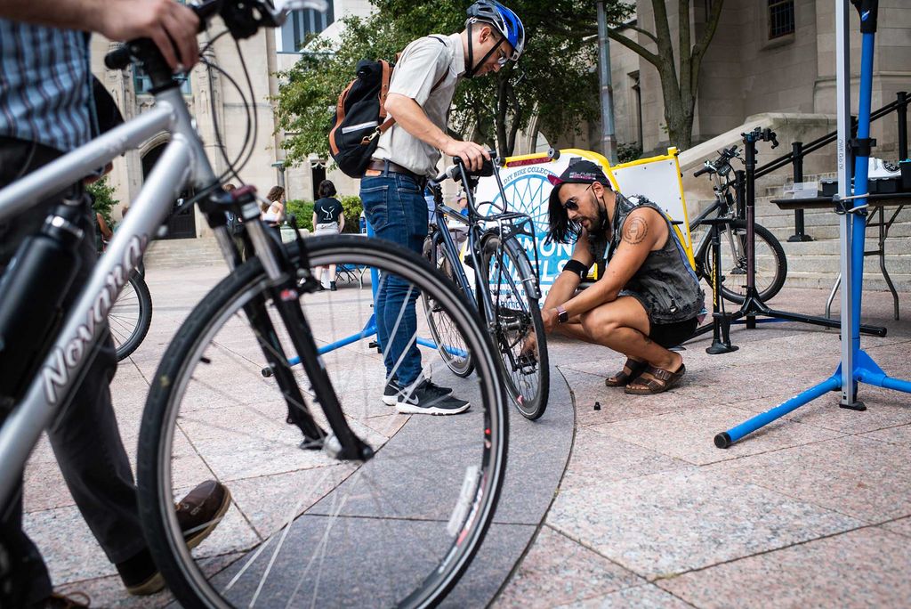 Photo of Teddy Heinrich, wearing a blue helmet and holding his bike, while Tristan Djaafar, crouching, in black, fixes something near his wheel. Another person holding their bike is seen in the left foreground. They appear to be on Marsh Plaza.