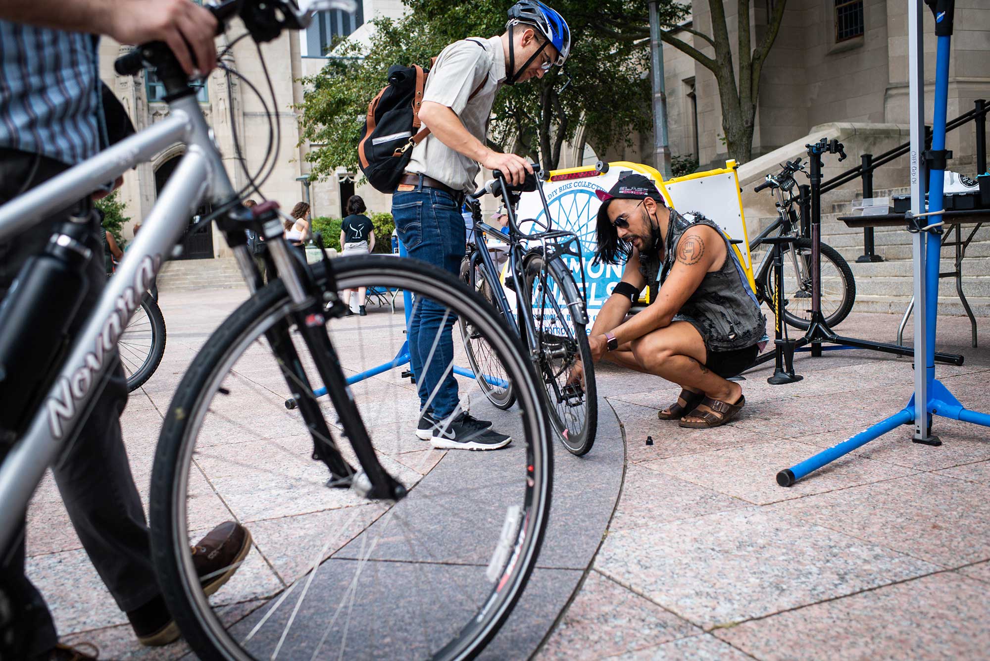 Photo of Teddy Heinrich, wearing a blue helmet and holding his bike, while Tristan Djaafar, crouching, in black, fixes something near his wheel. Another person holding their bike is seen in the left foreground. They appear to be on Marsh Plaza.
