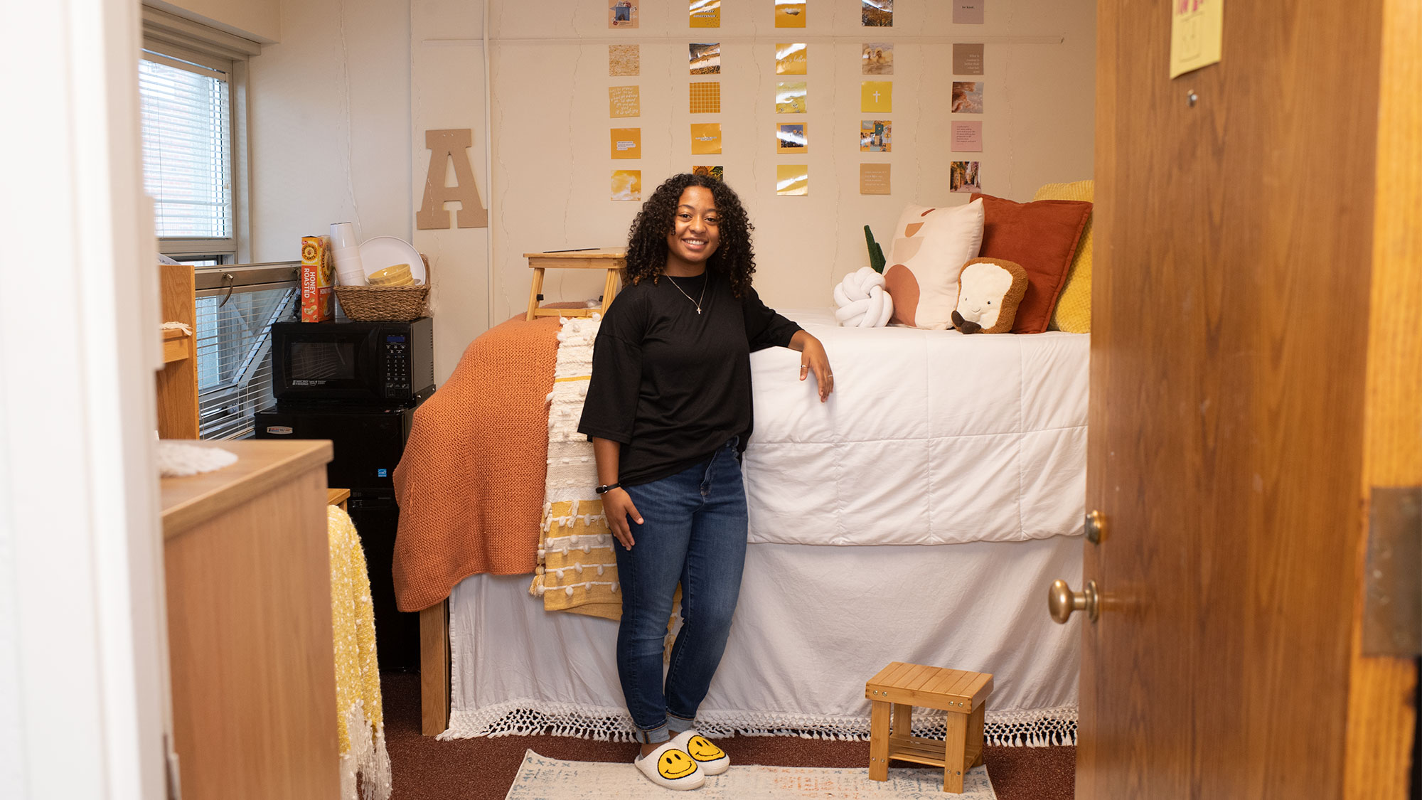 Photo of Anea Harris (Wheelock'24), in a black shirt, blue jeans, and slippers with smilie faces on them, resting her hand on her lofted bed in her dorm room. The walls are decorated with photos and the bed has pillows in the shape of a knot and a piece of toast.