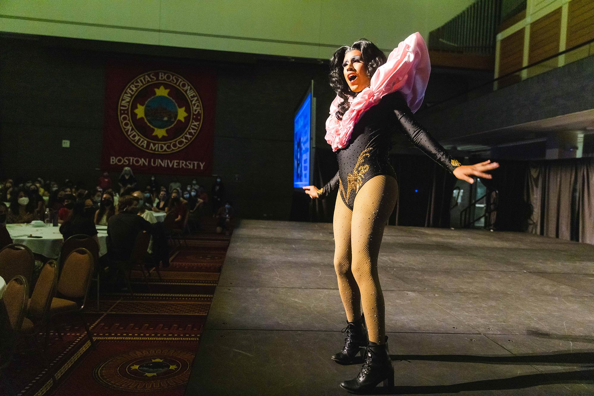 Photo of Linda Marie Póssa on stage, at Drag Bingo night at Metcalf Hall on Saturday night. Póssa wears a black and gold leotard, sparkling fish nets, black boot heels, and pink ruffles around their neck. They appear to be dancing or singing and they have black curly hair. Masked students are seated at white table cloth tables at left.