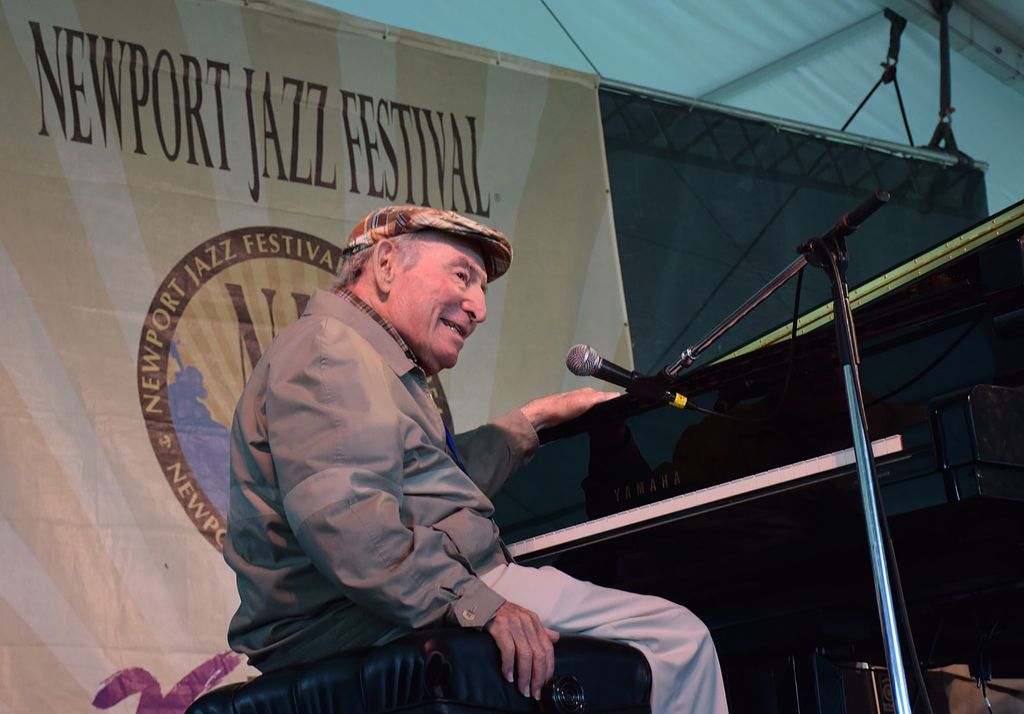 Photo of George Wein from 2014, wearing a plaid scally cap, and sitting at a piano. He smiles, and looks like he just finished playing. Behind him is a sign that says “Newport Jazz Festival.”