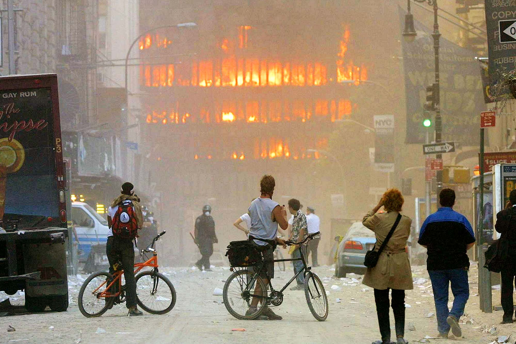 Photo of two people on bikes and other bystanders looking towards the twin towers as they burn. The air is filled with ash and the photo is very hazy.