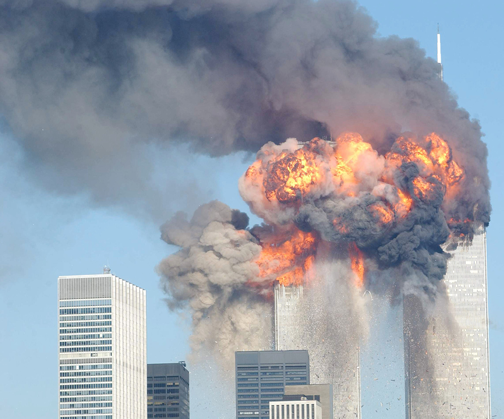 A photo of a fiery, orange blasts coming out of the World Trade Center after being hit by two planes September 11, 2001 in New York City. Huge clouds of smoke stretch into the blue sky.