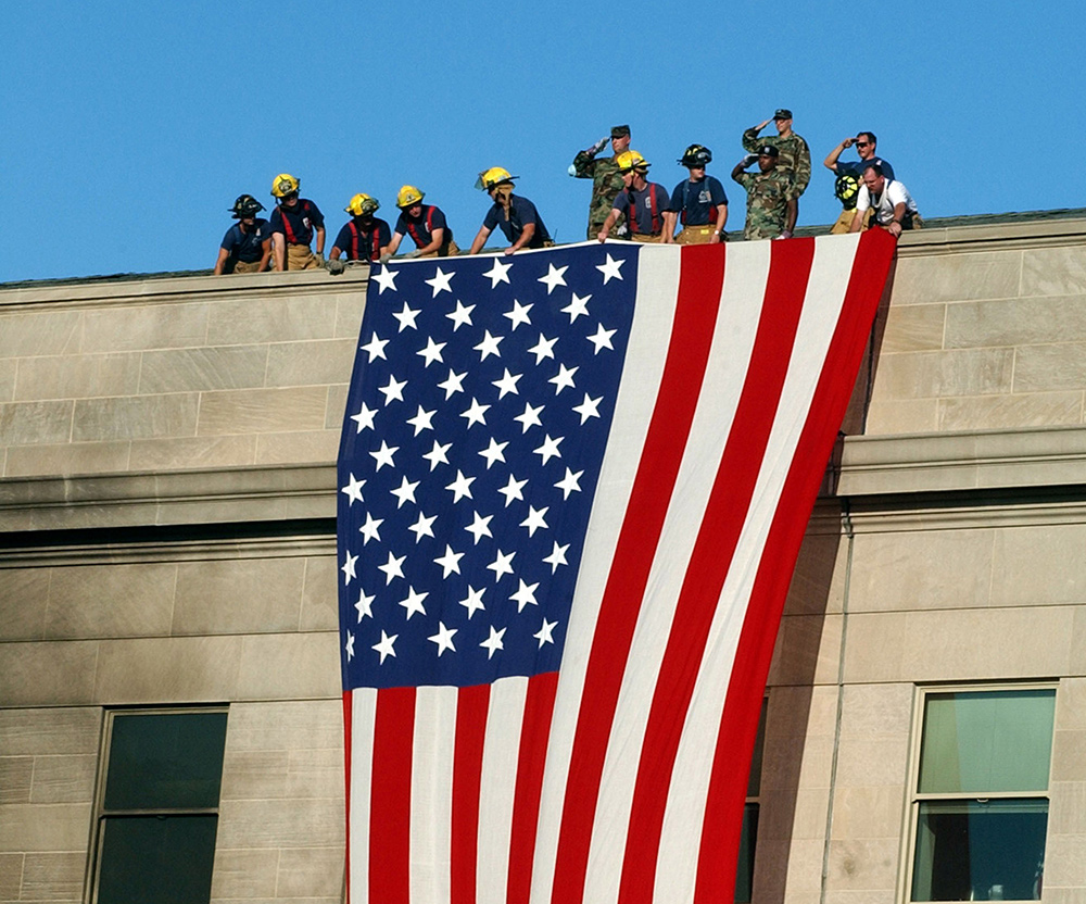 Photo of firefighters extending a giant American flag over the side of the pentagon after it was attacked. The sky behind them is bright blue.
