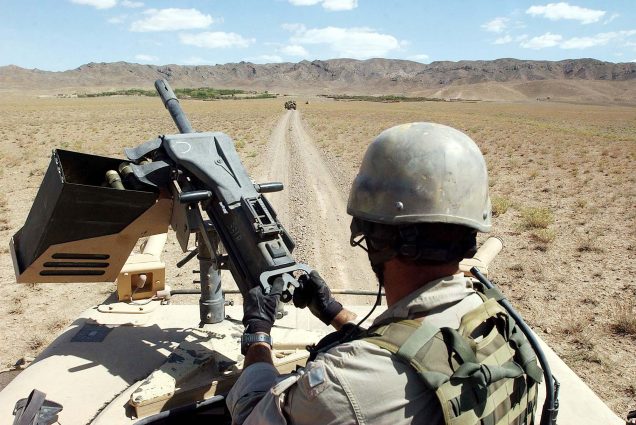 Photo of U.S. Special Forces soldier with Task Force 31 manning a gun on the way to conduct joint village searches with the Afghan National Army March 29, 2004 in southeast Afghanistan. He wears camouflage combat gear and holds a machine gun. In the background, a dusty road and mountains are seen.