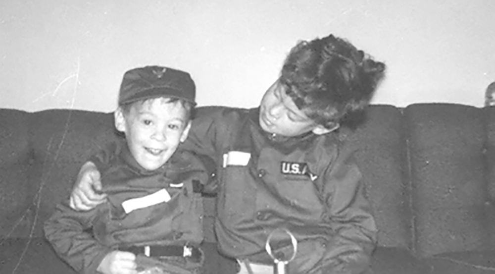 Black and white photo of Jay and Glenn as young boys. One has their arm around the other, and the boys smile. They wear jackets that say US Army on them.