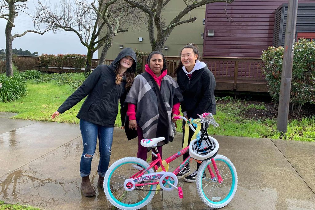Photo of Angela Tsai and two other community members who pose with a brightly-colored kids' bike. They all wear jackets and smile.