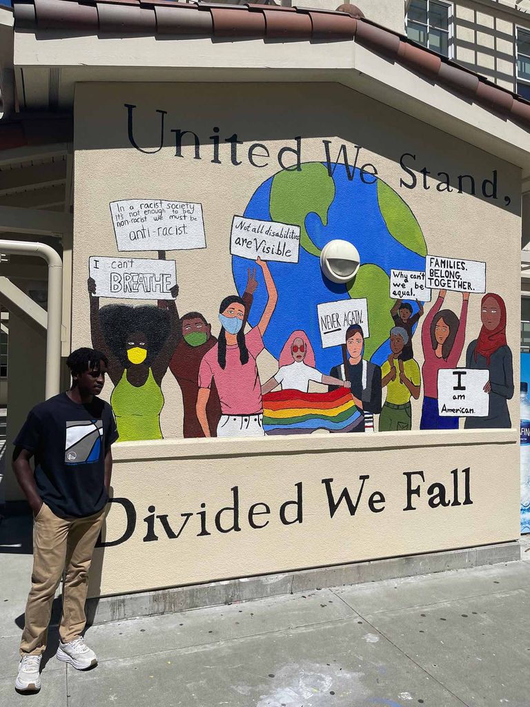 Photo of Tyler Johnson standing in front of a mural he helped design after someone spray painted hate speech on this wall at his high school. Johnson stands in a black t-shirt and khaki pants at left. Behind him, the mural has a tan background and reads “United we stand, divided we fall” and features a diverse group of people standing in front of the earth, with signs that say “I can’t breathe,” “Not all disabilities are visible,” “never again!” “families belong together!,” “I am American!” and others.