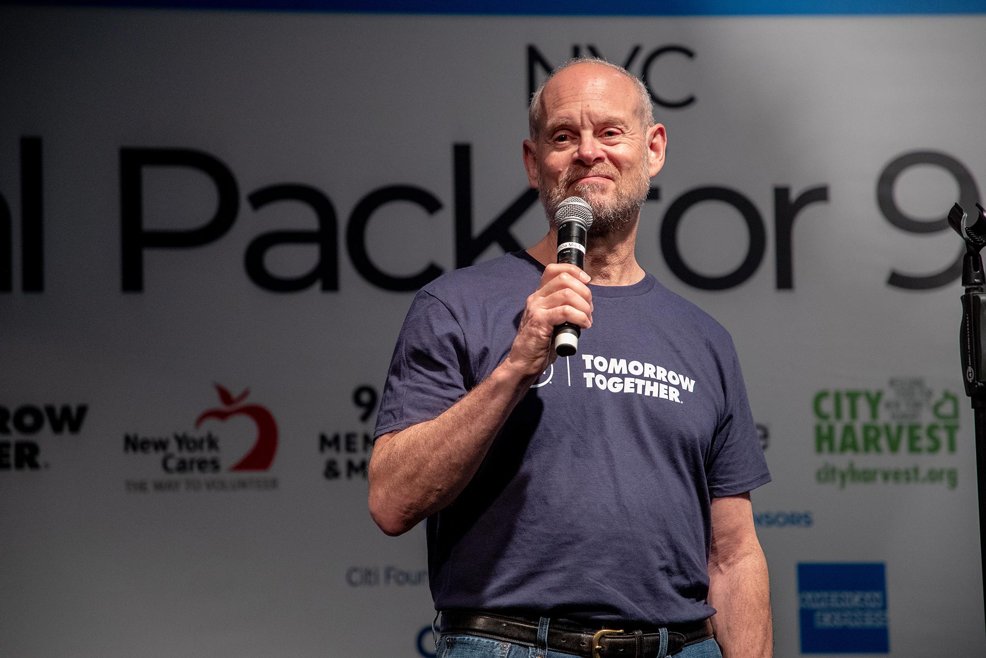 Photo of Jay Winuk, at an event that passed out meals to people in need in 2019, speaking into a microphone. He wears a grayish blue t-shirt that reads “Tomorrow together”