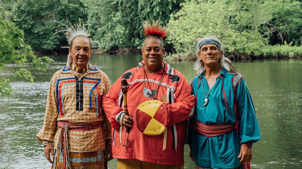 Photo of three members of the group Eastern Medicine Singers, who are an Algonquin troupe from Rhode Island that honor Indigenous voices. Three mid to late-aged. They wear indigenous shirts, sashes, and head pieces, and the man in the center holds what looks like a drum.