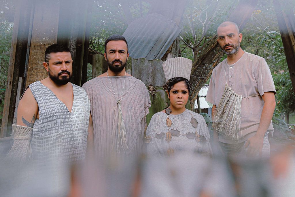 Photo of four members of the band Combo Chimbita, an avant garde guacharaca band, wearing flowing gray outfits. The photo is blurred at the top and bottom and the band looks serious. 