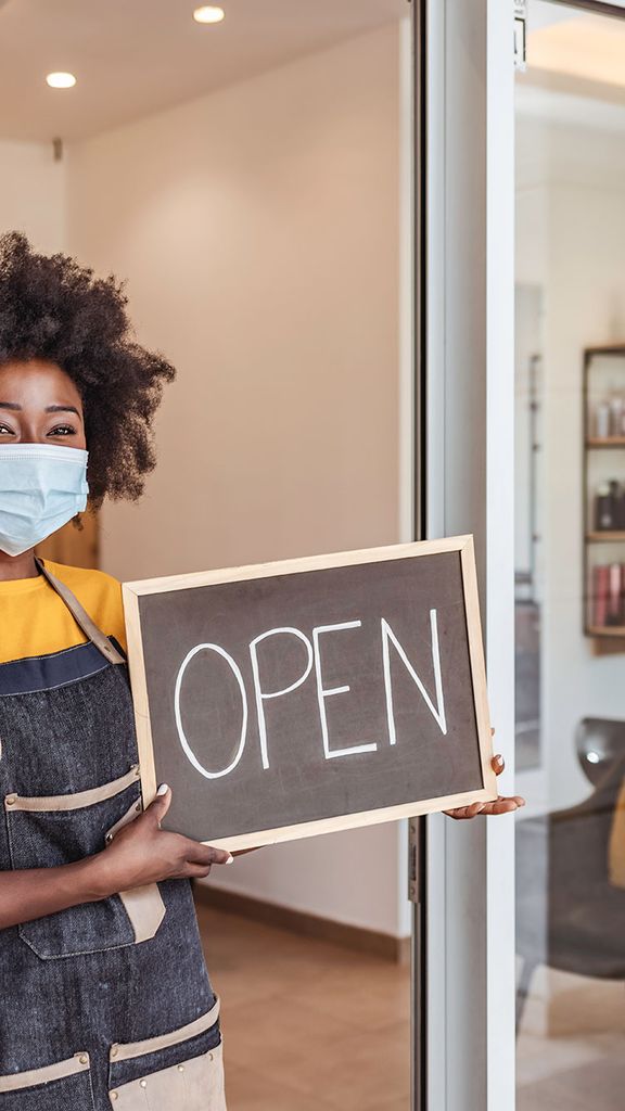 Photo of a black woman entrepreneur holding an chalkboard open sign at the front of her business. She wears a face mask and a stylish apron.