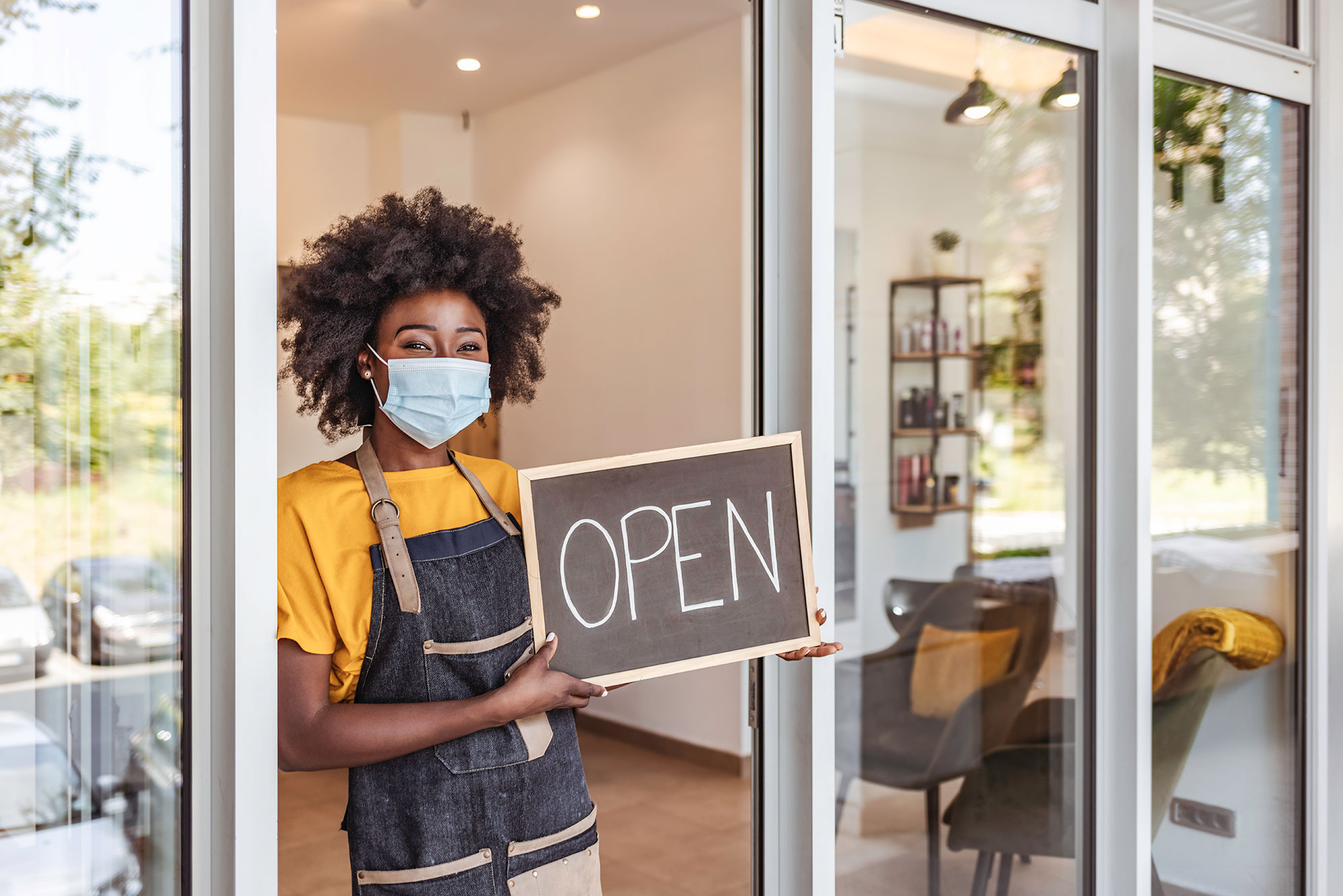 Photo of a black woman entrepreneur holding an chalkboard open sign at the front of her business. She wears a face mask and a stylish apron.