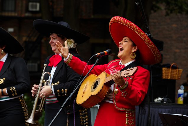 Photo of Veronica Robles, dressed in a red mariachi outfit with gold trim, and a wide brimmed red sombrero singing joyfully into a microphone and holding a 5 string acoustic guitar. Other members of the band, dress in black, and holding horns, are seen behind her.