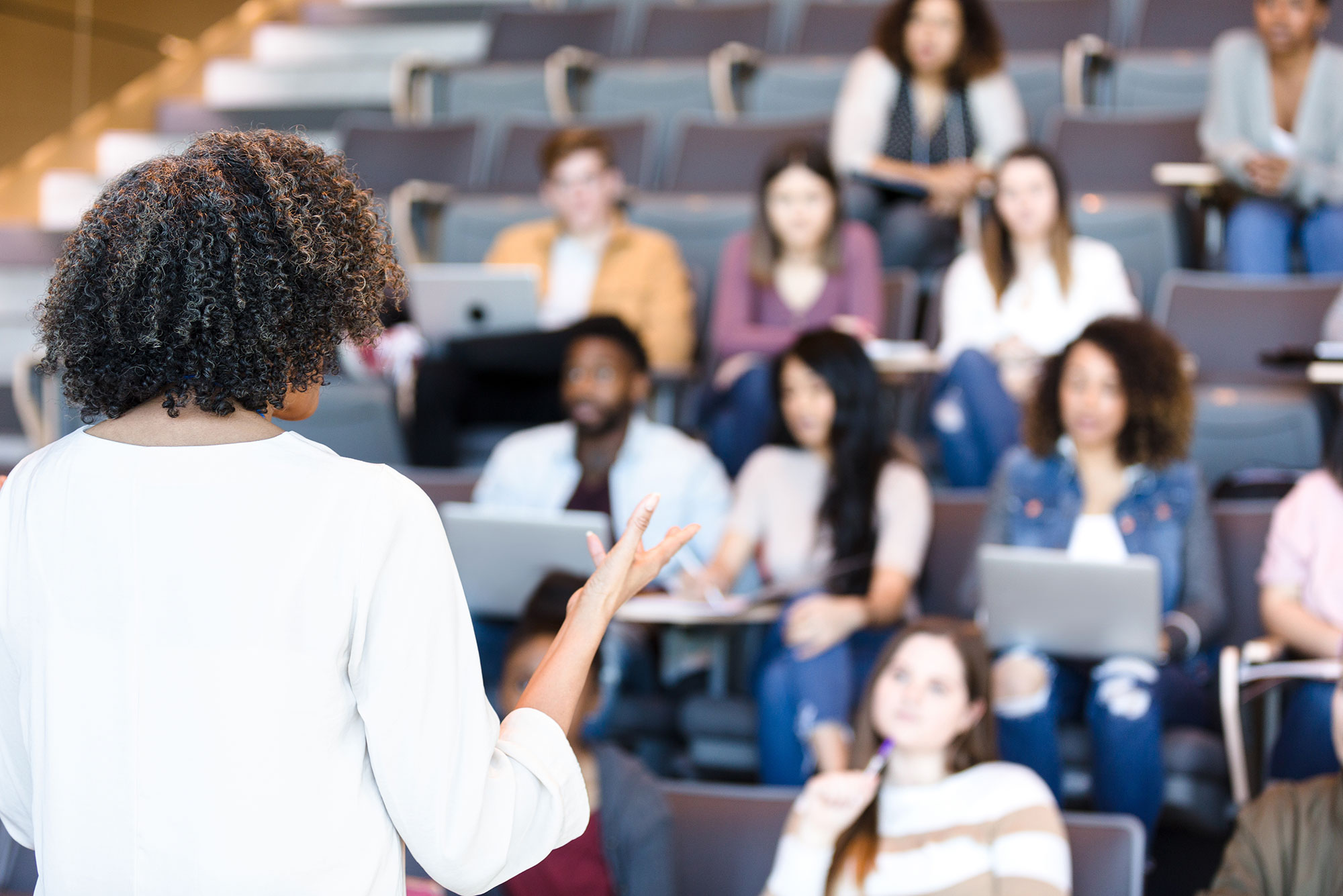 Photo of a youngish, Black woman, with curly almost shoulder length hair, wearing a white shirt, teaching at the front of a lecture hall. She is seen from behind.