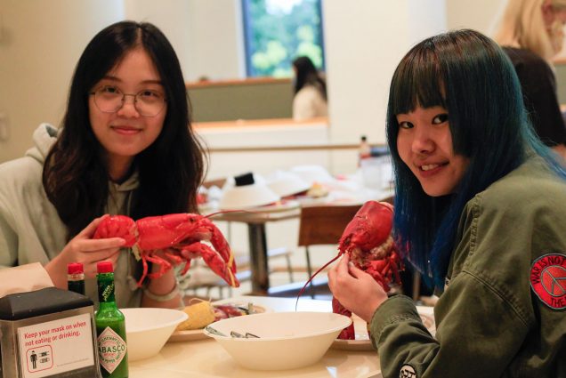 Photo of Yan Huang QST'22 and Zitong Zhao QST'22 holding their lobsters before eating them. They are sitting in Marciano Commons and there are a few bowls on the table.