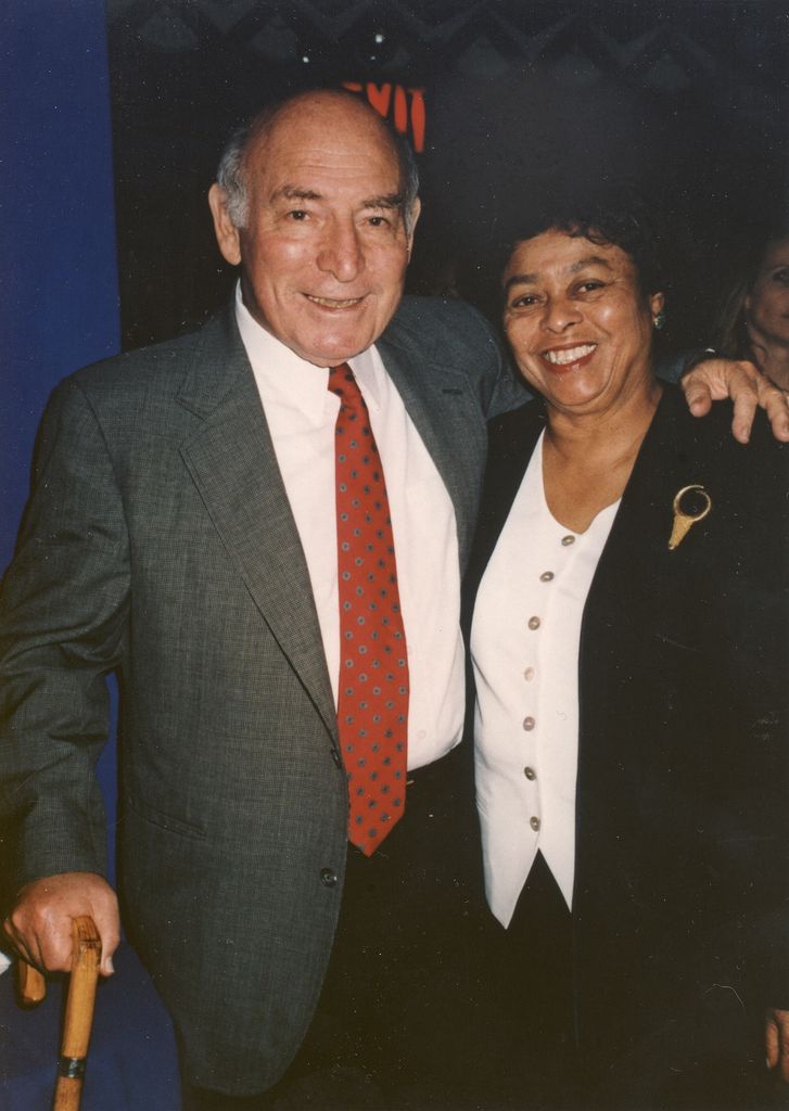 Photo of George Wein, his wife Joyce, undated. George wears a suit, red tie and has his left arm around Joyce. The couple looks to be about 60 or 70 years old. Wein holds a cane in his right hand. Joyce wears a black sweater and white blouse.