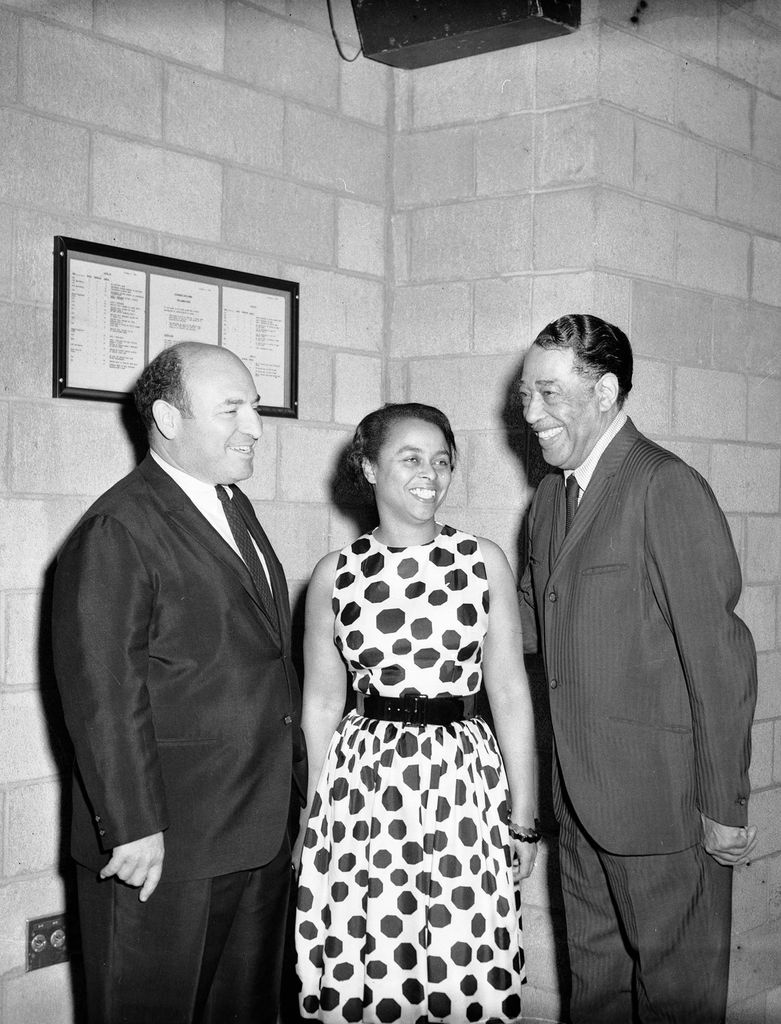 Black and white photo of George Wein, his wife Joyce, who wears wearing polka dot dress, and Duke Ellington, smiling, at Pittsburgh Jazz Festival in Civic Arena, Pittsburgh, Pennsylvania, 1965. The men wear dark suits and they stand in a building with brick walls.