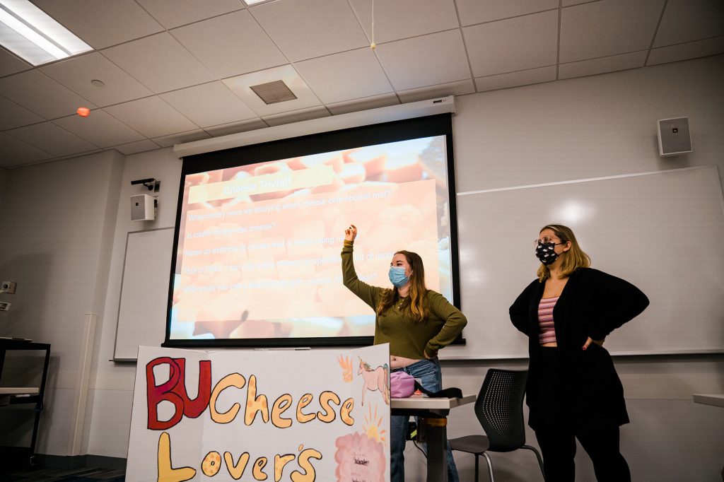a photo of BU Cheese Society president Hannah Gorman (Sargent’22) (left) and vice president Kimberly Candelario (CAS'22) leading the first meeting of the semester. They stand in front of a projector screen and next to a large, handmade sign that reads "BU Cheese Lovers"