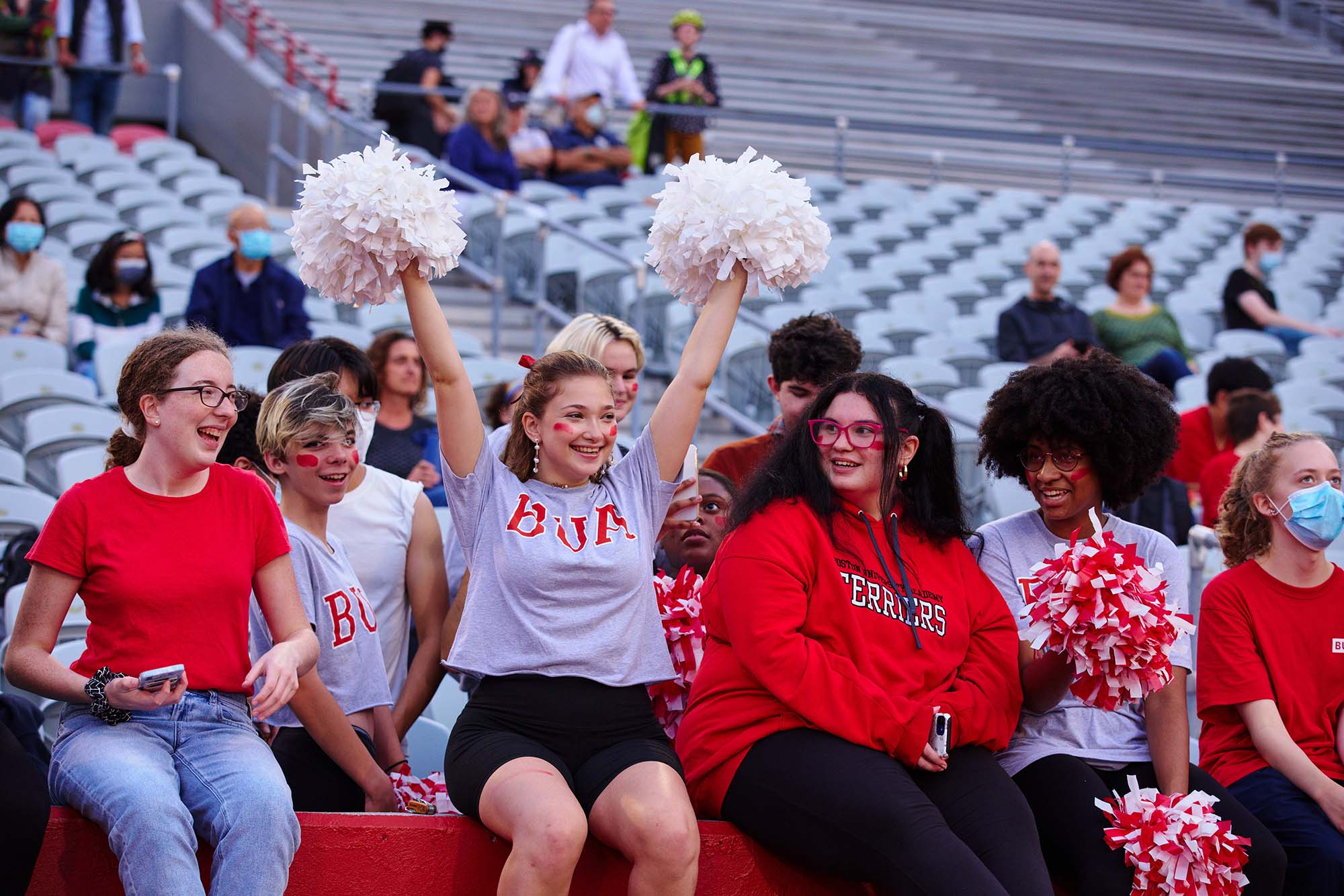 Photo of Boston University Academy fans, from left, Julia Dickinson (BUA’22), Quinn Lynn (BUA’25), Claudia Woolf (BUA’22), Suzie Marcus (BUA’22), Amaya Willis (BUA’22), and Ivy Keldsen (BUA’23), cheering on the BUA girls soccer team in the stands at Nickerson Field. Claudia waves white pom poms as her friends around her look on.