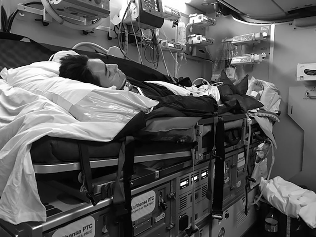 Black and white Photo of Ethan Wang on a lufthansa flight in an emergency Patient Transport unit. He lays down on a stretcher, and there are lots of wires and medical equipment around him.