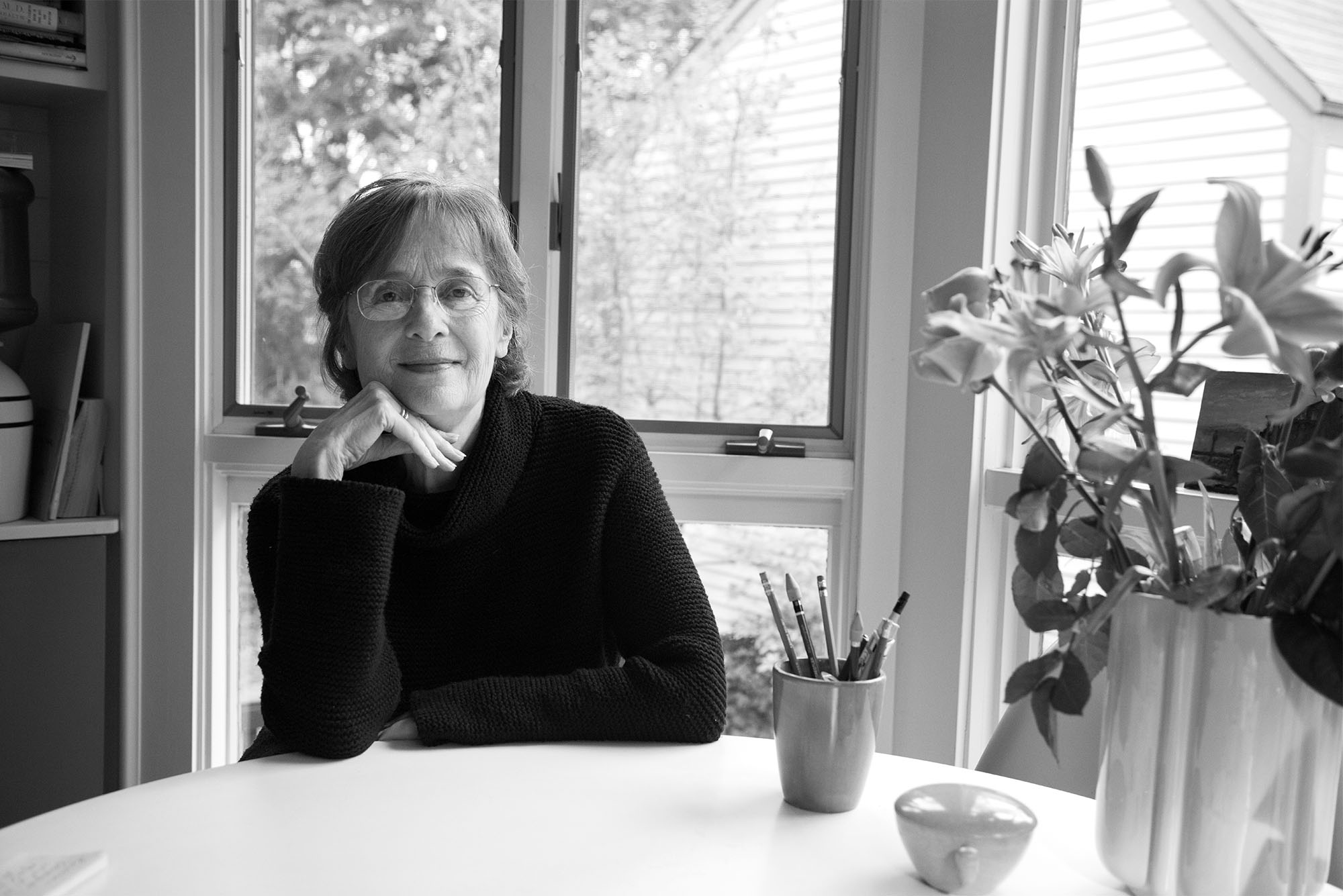 a black and white photo of author Gail Mazur sitting at table with chin resting on her hand in front of a bright window.