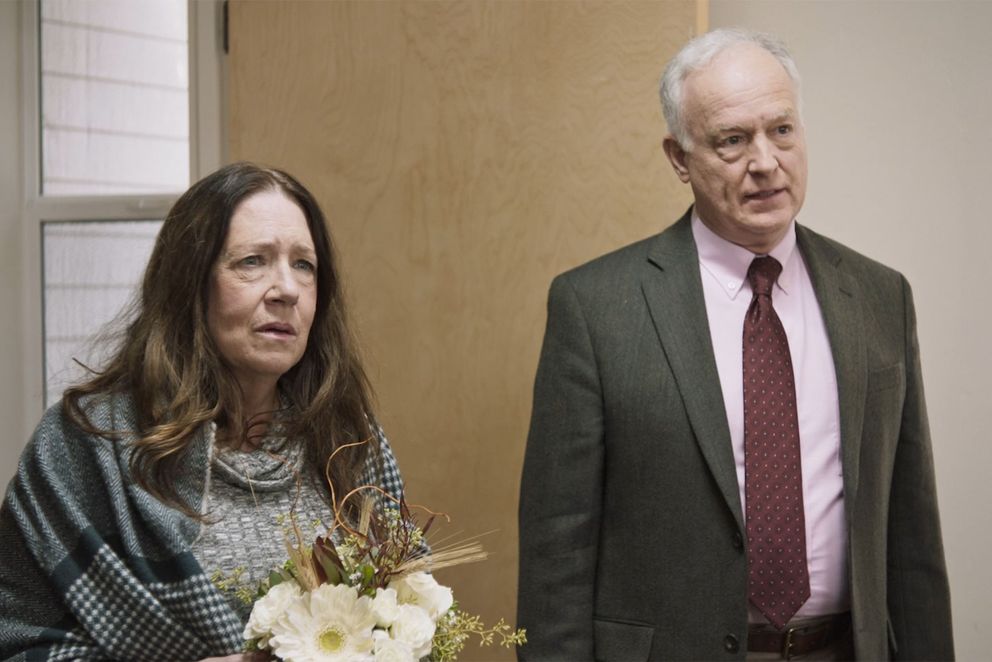 screenshot from new film Mass: (from left to right) Both looking distressed, Ann Dowd holds a bouquet and stands next to Reed Birney in hospital doorway