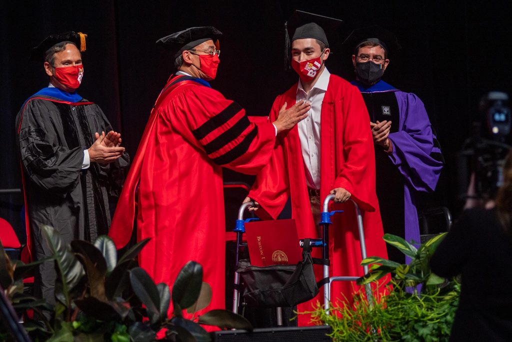 Photo of Ethan Wang on stage with his dad Willis. They are both dressed in graduation caps and gowns, and wear red face mask. Ethan uses his walker on stage; in the walker’s pocket is his red diploma.