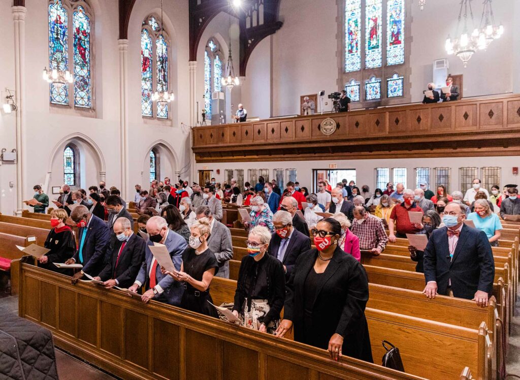 Photo of people standing in the pews at the 2020 Boston University Baccalaureate Ceremony at Marsh Chapel on October 3, 2021. They all wear face masks, and the chapel's large stain glass windows are seen. 