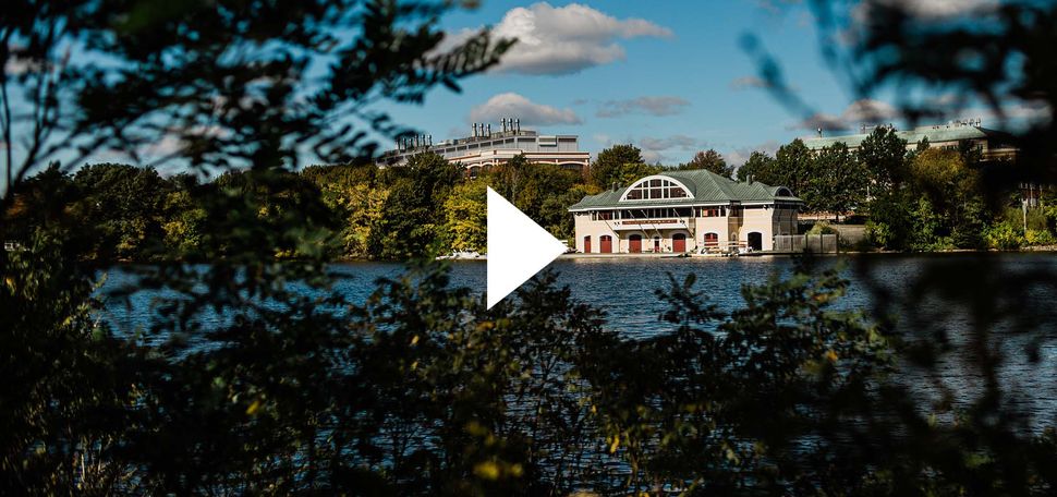 Photo of DeWolfe Boathouse taken from across the Charles River on a sunny fall day. Small clouds are seen in a brilliant, blue sky. The Boathouse is tan with a greenish gray roof. Play button is overlaid.