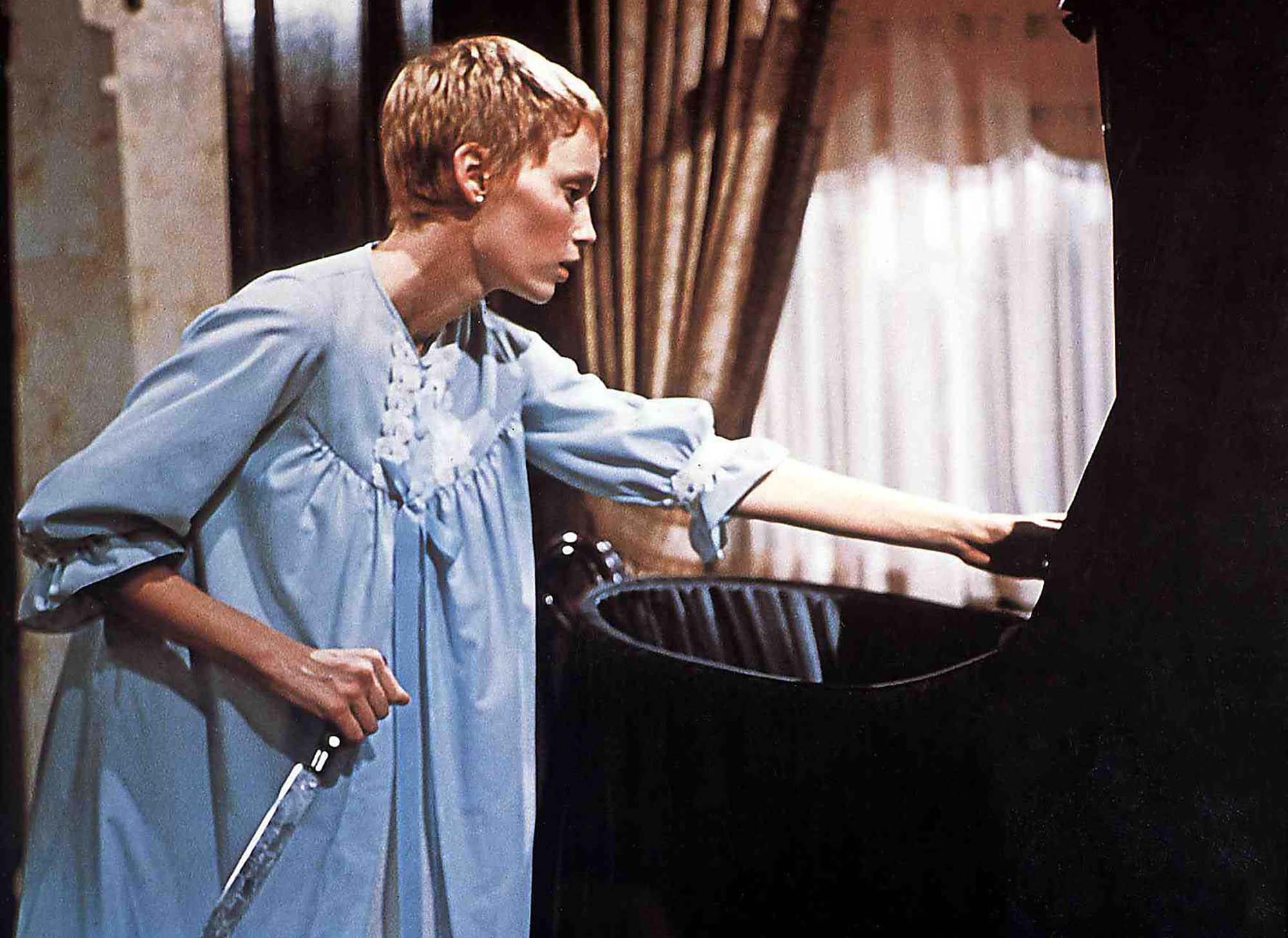 Still from the horror film Rosemary's baby. Rosemary stands in a blue night gown with a knife in her hand over a black baby cradle.