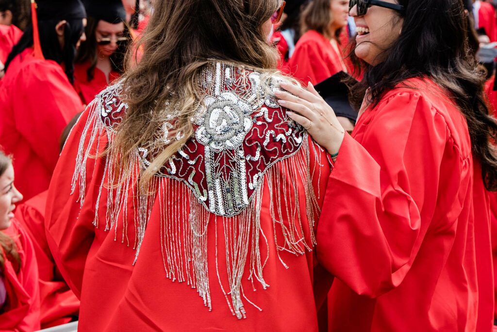 Two graduating student in graduation gowns laugh. One student is wearing a shawl over their graduation gown.