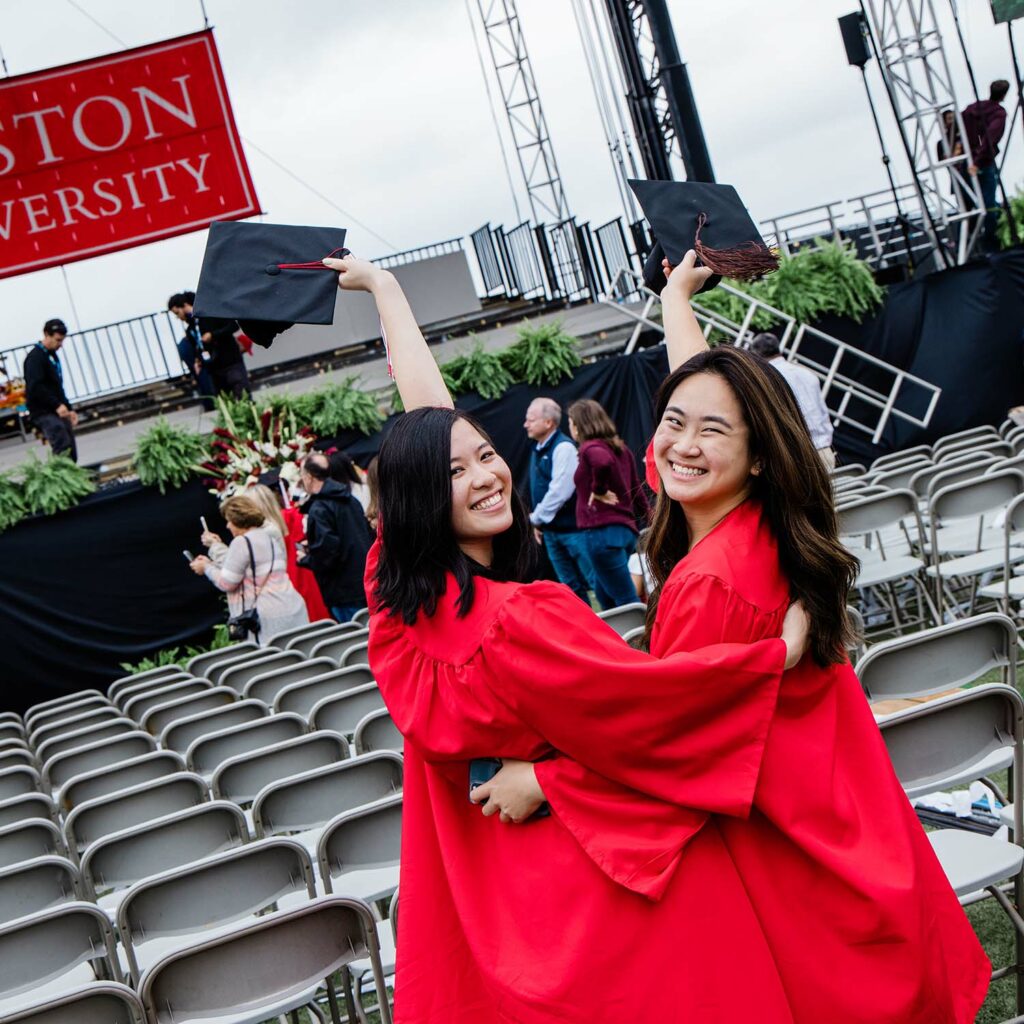 Two students hold their graduation caps in the air and look over their shoulders smiling for a photo.